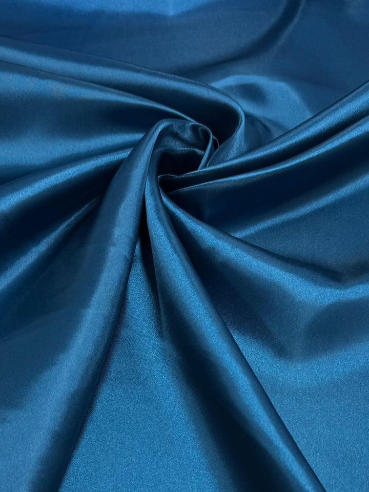 Super Cheap Fabrics' Satin - Teal - 150cm features smooth, glossy teal fabric draped into a central swirl, showcasing its rich color and sheen—ideal for creating elegant dresses.