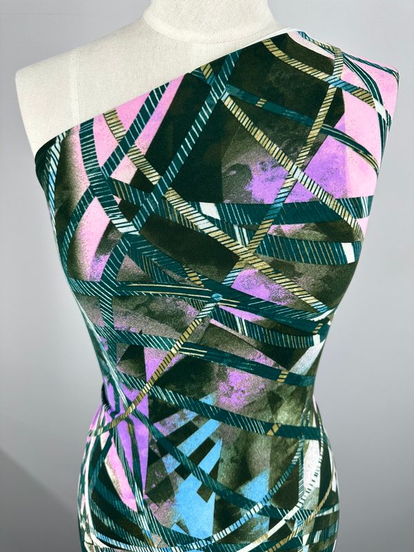 A mannequin showcases the "Printed Lycra - Tied Up - 150cm" dress by Super Cheap Fabrics, featuring a vibrant geometric pattern. The medium-weight fabric, made from Polyester/Spandex, highlights overlapping strips in shades of purple, pink, green, and teal against a white background for a modern and striking design.