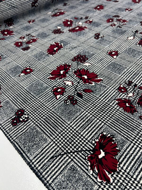 A close-up photograph of the Scuba Crepe - Hounds Dream fabric from Super Cheap Fabrics. The material, textured and lightweight, features red flowers scattered across the checkered black and white design, adding a vibrant contrast to the monochrome background. The texture is clearly visible, highlighting the intricate details of this 160cm wide fabric.