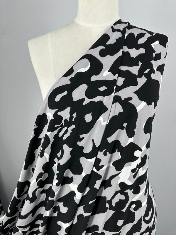 A mannequin is draped with Super Cheap Fabrics' medium-weight Printed Lycra - Camo, which features a contemporary camouflage motif of irregular black shapes on a light background. The fabric measures 150cm in width and appears soft and slightly stretchy, making it suitable for clothing or upholstery.