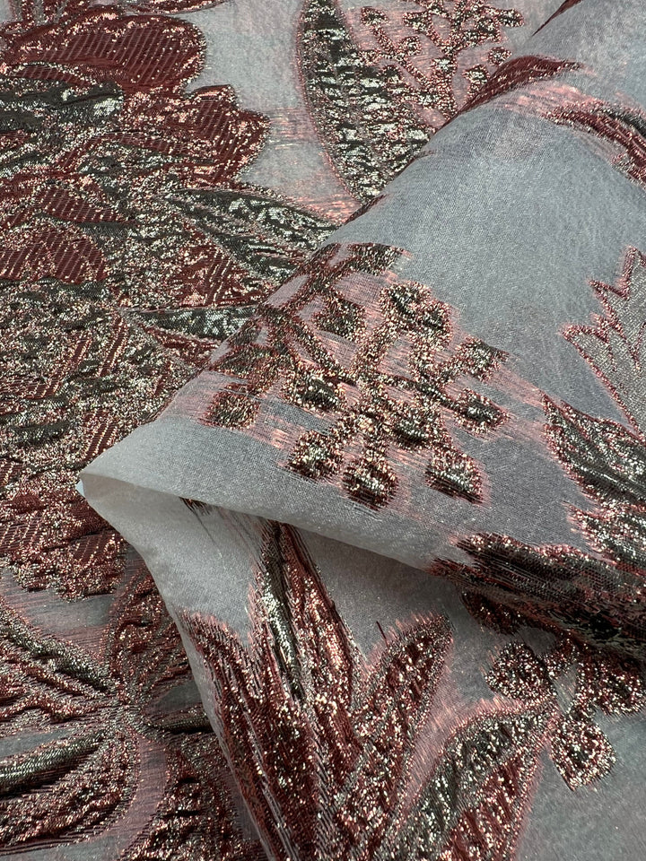 Close-up of the Designer Metallic Brocade - Anne - 160cm from Super Cheap Fabrics, featuring an intricate floral pattern. The design showcases shimmering rose gold and metallic green threads against a translucent, polyamide light gray background. The texture appears rich and detailed, with the lightweight brocade fabric gently folded to display different angles.
