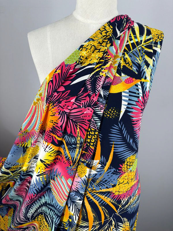 A mannequin draped in beautiful prints on vibrant fabric showcasing a colorful, tropical pattern. The design features an assortment of leaves and foliage in bright shades of pink, yellow, green, blue, and white on a dark navy background. This stunning display is made from Super Cheap Fabrics' Printed Lycra - Jumba - 150cm.