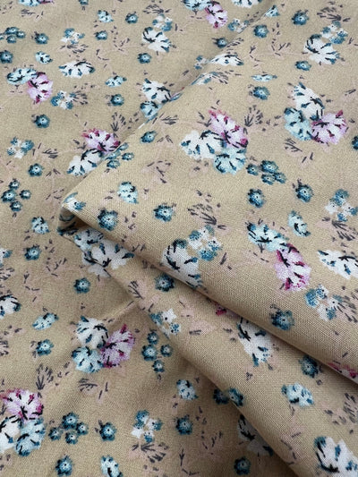 A close-up view of Super Cheap Fabrics' Printed Rayon - Pansy - 145cm, featuring a delicate floral pattern in shades of blue, purple, and white on a beige background. This versatile fabric is shown folded, revealing its vibrant prints on both visible layers.