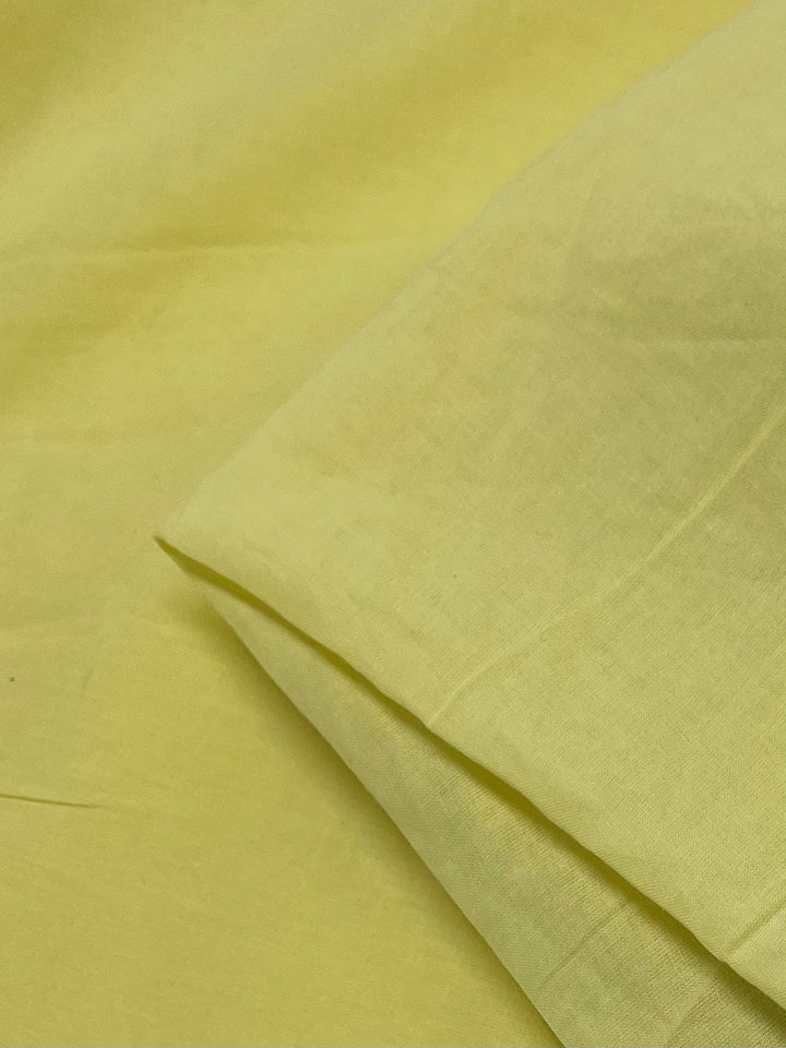 A close-up image of a yellow cotton fabric, showing a smooth and slightly wrinkled texture. The fabric appears to be folded in the upper right corner, highlighting its extra lightweight nature. The color is a bright, cheerful yellow with a hint of lima bean hue. This is the Cotton Voile - Lima Bean - 140cm from Super Cheap Fabrics.