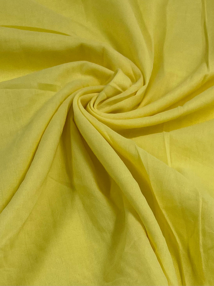 A close-up image of Super Cheap Fabrics' Cotton Voile - Lima Bean - 140cm, an extra lightweight, bright yellow cotton fabric twisted softly at the center, creating a spiral pattern. The material appears smooth, with gentle folds radiating outward from the central twist.