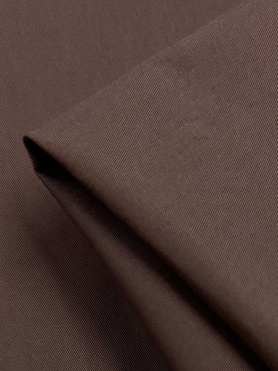 A close-up image of Super Cheap Fabrics’ Plain Sateen - Fondue Fudge, showcasing its 150cm wide brown cotton fabric with a diagonal weave pattern. The medium weight fabric appears folded, displaying its texture and smooth finish.