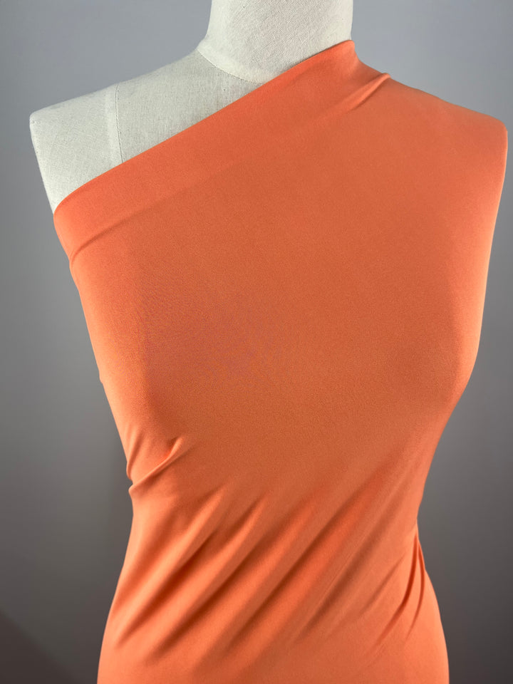 A mannequin dressed in a one-shoulder, form-fitting orange top made of 150 cm Super Cheap Fabrics' Plain Lycra in Sunkist is set against a plain gray background. The fabric drapes smoothly over the bust and torso, creating gentle folds and subtle shadows.