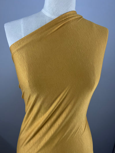 A mannequin is draped in an environmentally responsible, golden-yellow, one-shoulder fabric with a subtle texture. The material appears smooth and slightly shiny, creating gentle folds and curves along the mannequin's form. The background is plain and gray. The fabric used is Bamboo Jersey - Arrowwood - 160cm by Super Cheap Fabrics.