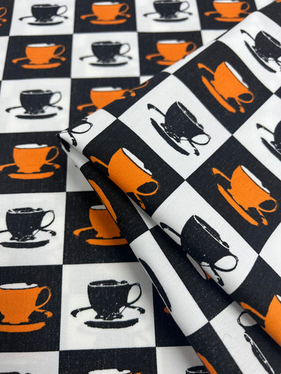 This 100% Cotton Drill - Orange MugKnight - 155cm from Super Cheap Fabrics features a checkered pattern with orange and black coffee cups on alternating black and white squares. The design's lightweight texture showcases repeated images of the coffee cup motif, creating a visually striking contrast.