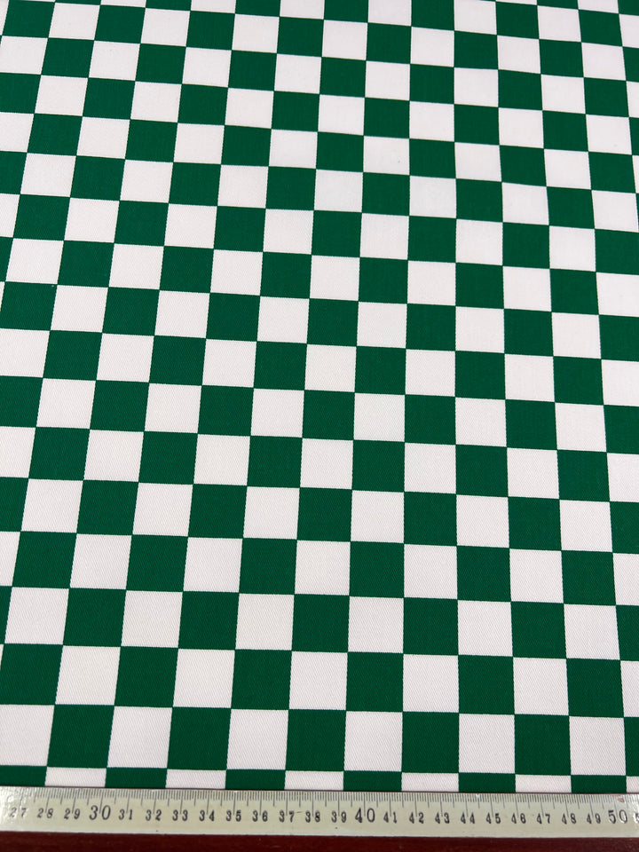 A piece of Super Cheap Fabrics' Cotton Drill - Green Chess - 155cm with a green and white checkerboard pattern is laid out flat. A ruler placed at the bottom edge measures the fabric, showing numbers from 25 to 45 centimeters. The checkered squares are evenly sized, highlighting the fabric's durable diagonal weave.