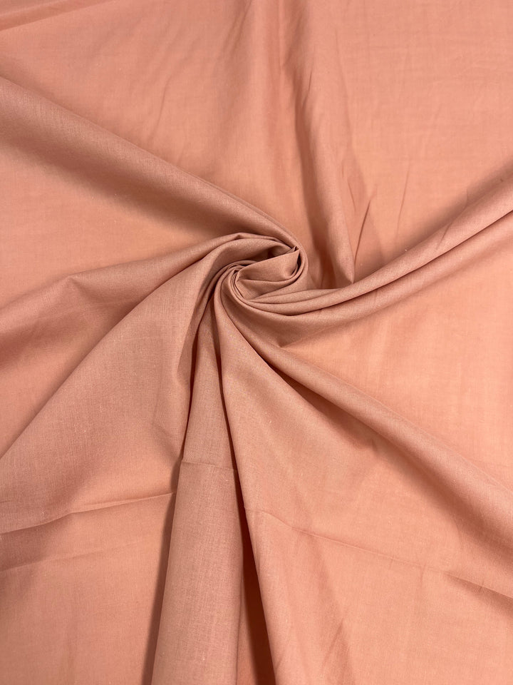 A close-up image of light pink or peach-colored fabric, made from 100% cotton, slightly wrinkled and gathered at the center into a twisted swirl pattern, creating a radial design effect around the twist. This extra lightweight fabric is perfect for children's clothing. The fabric shown is "Cotton Voile - Canyon Clay - 140cm" by "Super Cheap Fabrics.