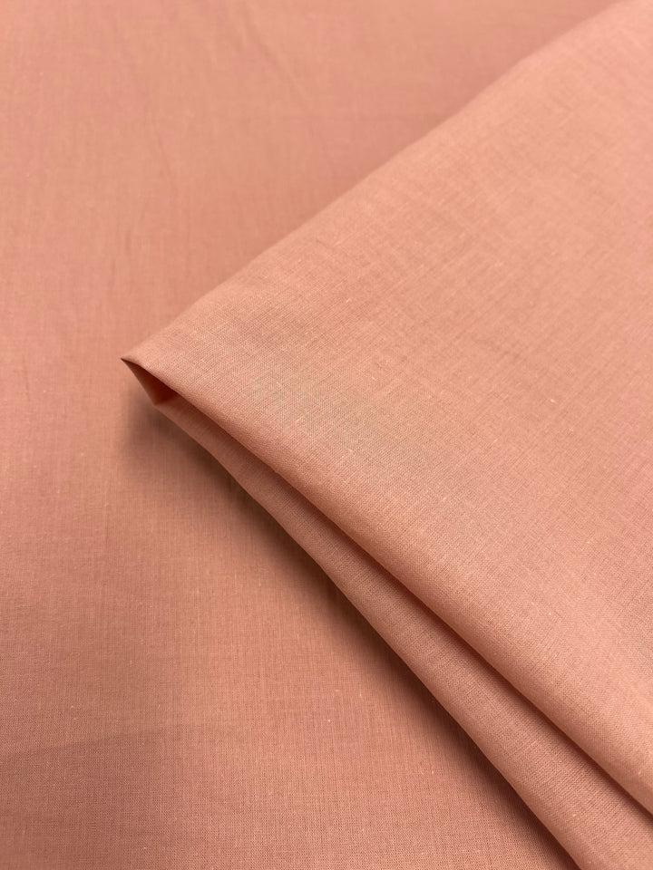 A close-up image of a folded piece of soft pink, 100% cotton fabric. The extra light weight material appears smooth and lightweight, with subtle texture visible on its surface. The fabric has clean edges and is neatly arranged on a flat surface, ideal for children's clothing. This is the Cotton Voile - Canyon Clay - 140cm from Super Cheap Fabrics.