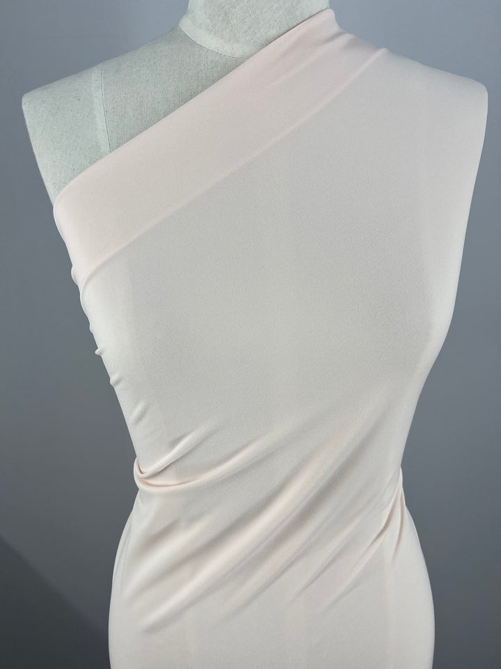 Close-up of a mannequin dressed in an asymmetrical, medium weight, one-shoulder dress. The Super Cheap Fabrics ITY Knit - Baby Pink - 150cm fabric is smooth and drapes elegantly with subtle ruching on the side, creating a sleek and refined look. The background is plain and gray, keeping the focus on the dress.