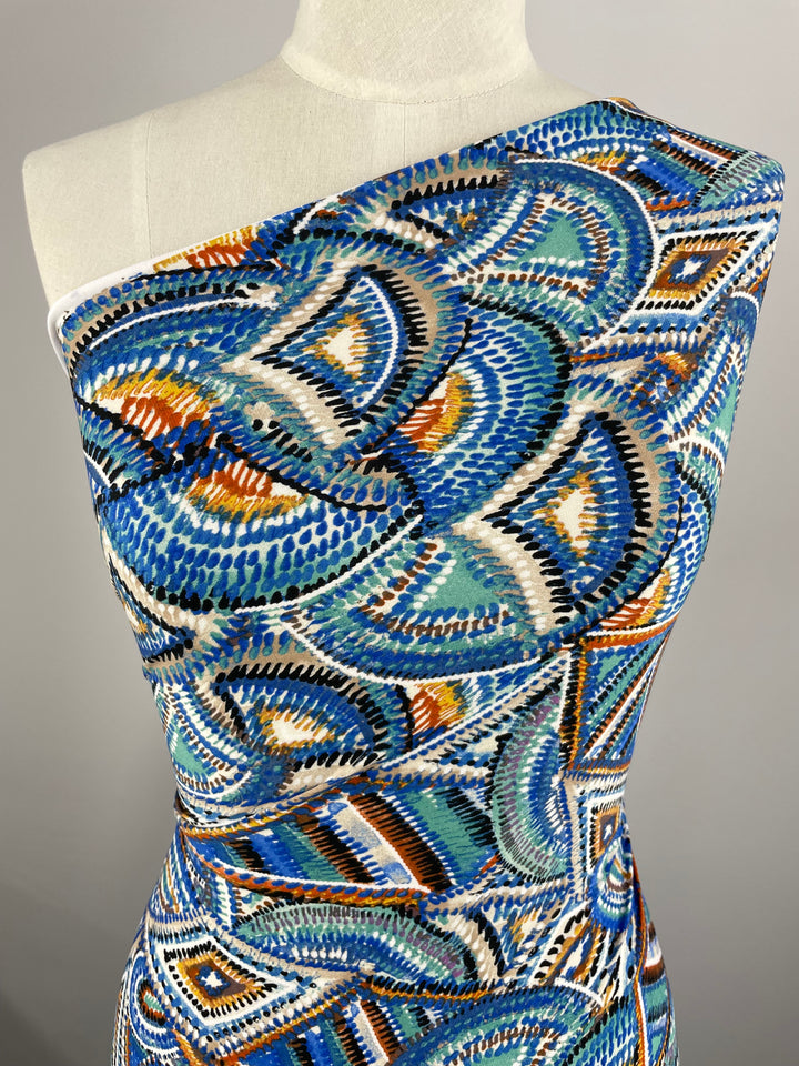 A mannequin is dressed in a fitted, colorful one-shoulder dress made from Super Cheap Fabrics' Printed Lycra - Mirage - 150cm. The medium-weight fabric showcases a swirling, abstract design with shades of blue, turquoise, orange, and white. The plain gray background highlights the vividness of the dress.