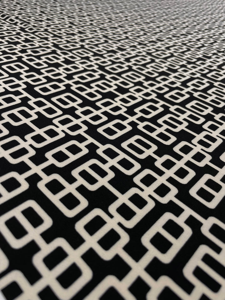 Close-up of a medium weight fabric with a geometric pattern of interlocking cream squares and rectangles on a black background. The Super Cheap Fabrics Printed Lycra - Link - 150cm design creates a visually intricate and repetitive motif, giving a modern and abstract appearance.