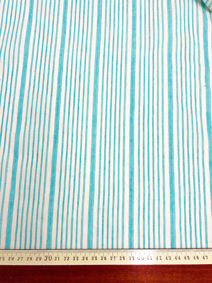 A piece of Cotton Voile - Light Blue- 150cm from Super Cheap Fabrics with thin, vertical teal stripes on a white background. A yellow measuring tape placed at the bottom indicates dimensions in centimeters, with markings visible from 27 to 36 cm. A small, partially visible rolled edge is at the top right corner.