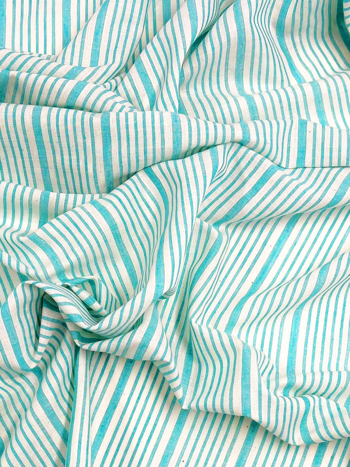 A close-up photo of **Super Cheap Fabrics Cotton Voile - Light Blue- 150cm** with a pattern of alternating vertical stripes in varying widths. The stripes, in shades of light blue and white, create a visually textured effect. The 100% cotton fabric appears loosely gathered or crumpled, showcasing its lightweight nature.