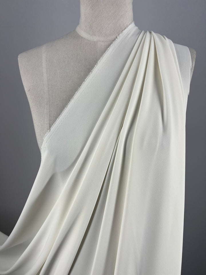 In an elegant display, the Double Knit Jersey - Ivory - 150cm from Super Cheap Fabrics is beautifully draped on a mannequin. The medium weight fabric flows smoothly over one shoulder, creating soft, cascading folds down the front. Its silky and light texture is perfectly highlighted against the gray background.