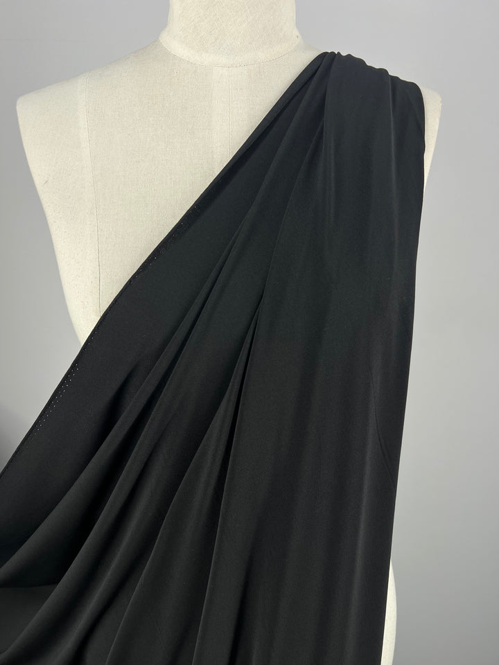 A close-up of a dress form draped with Super Cheap Fabrics' ITY Knit in Black, measuring 150cm. The medium weight fabric smoothly flows over the shoulder and drapes across the body, creating soft, elegant folds. The background is plain and gray, highlighting the sleek and simple design of the draped fabric.