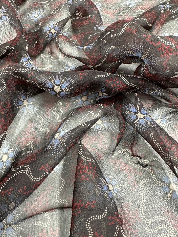 A piece of Pure Printed Silk - Artisan - 140cm from Super Cheap Fabrics draped in folds, featuring an intricate pattern. The design includes small white dots forming wavy lines, clusters of red floral shapes, and blue and white clustered flower motifs on a black and gray background, epitomizing high-end fashion.