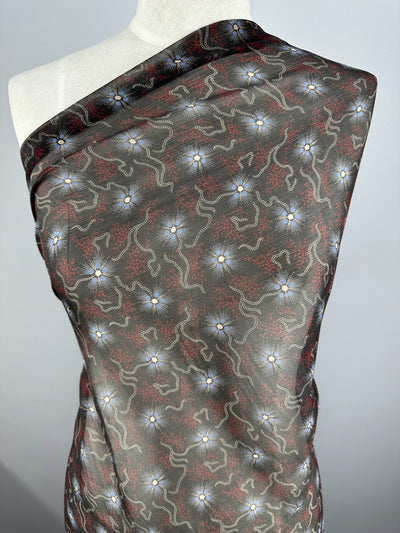 A mannequin draped in Super Cheap Fabrics' Pure Printed Silk - Artisan - 140cm showcases a dark fabric adorned with an intricate pattern of interconnected, flowing lines in red and light grey, complemented by small, white, flower-like shapes. The background is a gradient of black to dark brown with a subtle sheen, epitomizing high-end fashion.