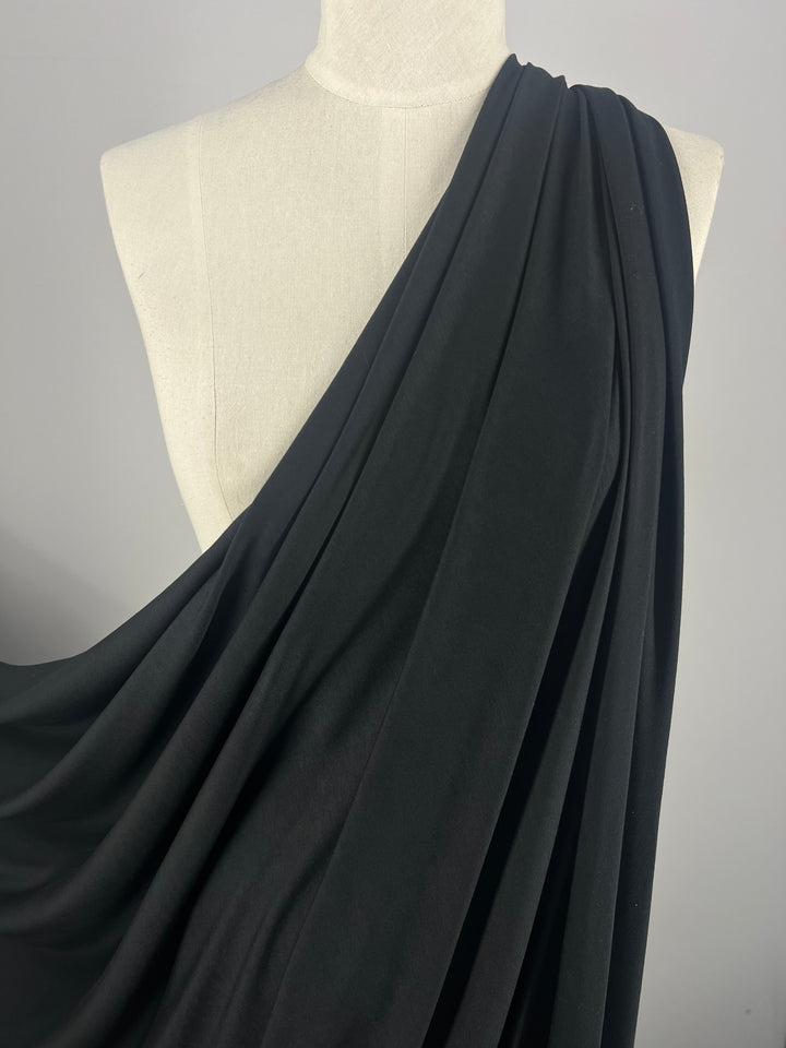 A white dress form is draped in a swath of dark, flowing Super Cheap Fabrics' Cotton Lycra - Black - 160cm, resembling a toga. The fabric is gathered and arranged over one shoulder, cascading down the front and side of the form, creating an elegant, classical appearance.