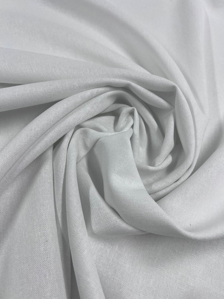 A close-up of the Super Cheap Fabrics' Linen Rayon Blend in white (137cm) elegantly folded into a spiral pattern, creating soft ridges and shadows. The fabric's smooth texture and slight sheen highlight its delicate drape.
