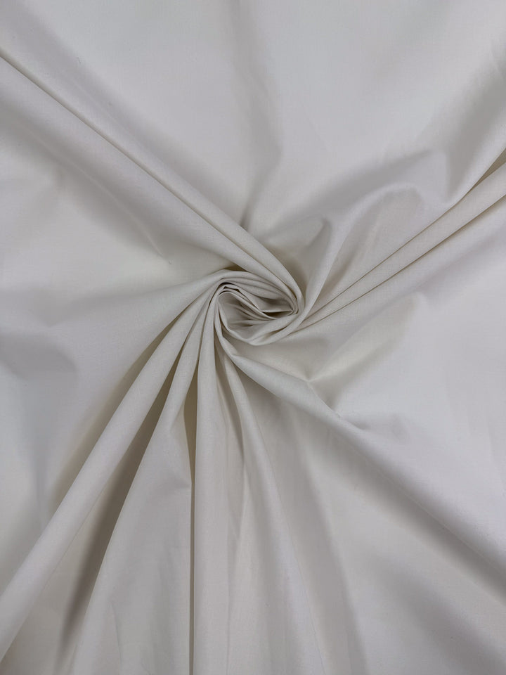 A close-up of Super Cheap Fabrics' Cotton Voile - Gardenia - 150cm shows light beige fabric twisted into a spiral at the center, creating soft folds and gentle shadows around the swirl.