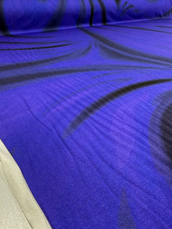 A close-up of Super Cheap Fabrics’ Viscose Knit - Ocean Blur (140cm), showing smooth, vibrant blue and purple stretch fabric with black abstract swirls and a small beige edge.