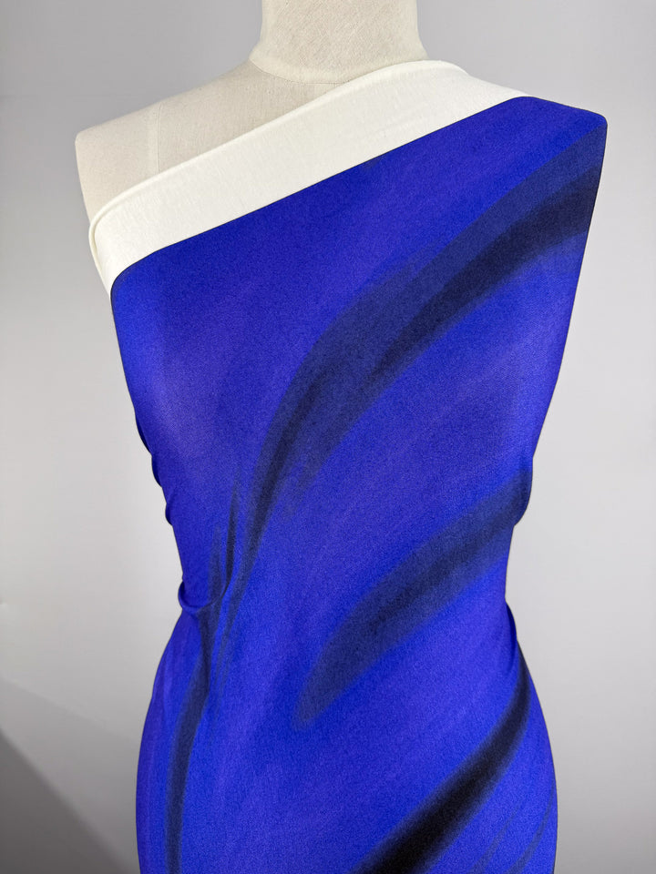 A mannequin displays Super Cheap Fabrics' Viscose Knit - Ocean Blur (140cm), a one-shoulder vibrant blue stretch viscose with black and dark blue brushstroke patterns and a white border, set against a plain background.