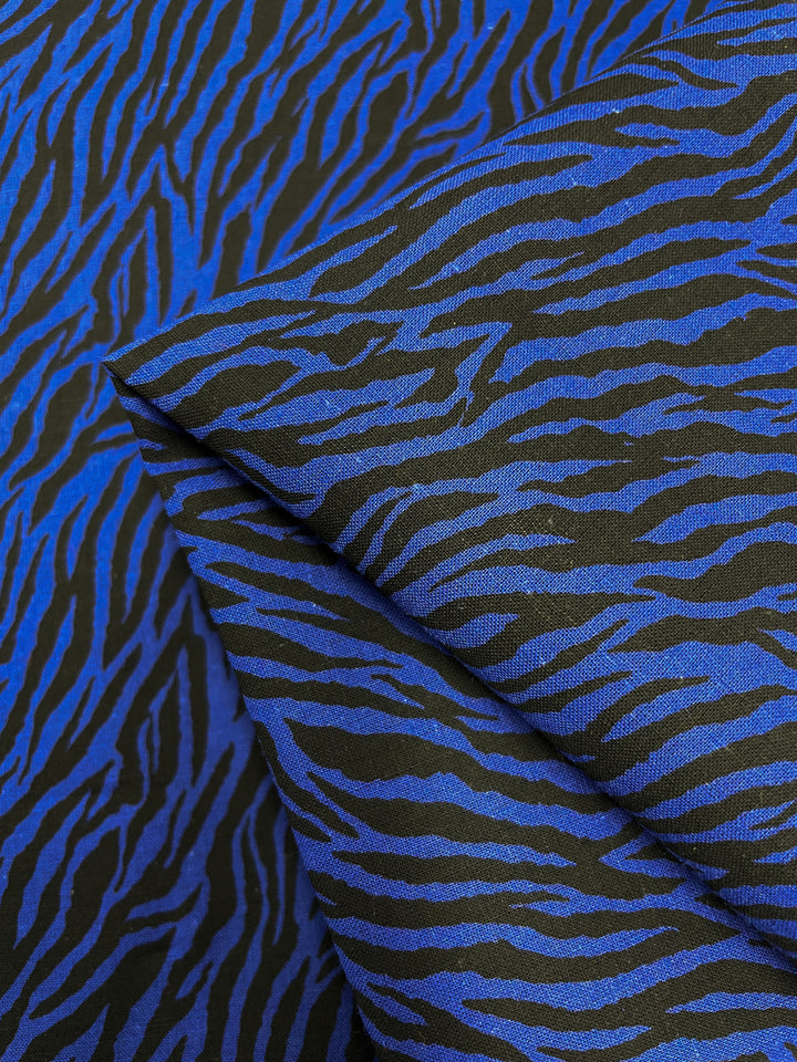 Close-up of the *Linen Voile - Cobalt Zebra - 140cm* from Super Cheap Fabrics, featuring a vivid blue and black zebra stripe pattern. The slightly folded Linen Voile showcases the intricate design's consistency. The bold colors and animal print create a striking visual effect, making it perfect for trendy home decor that also offers breathability.