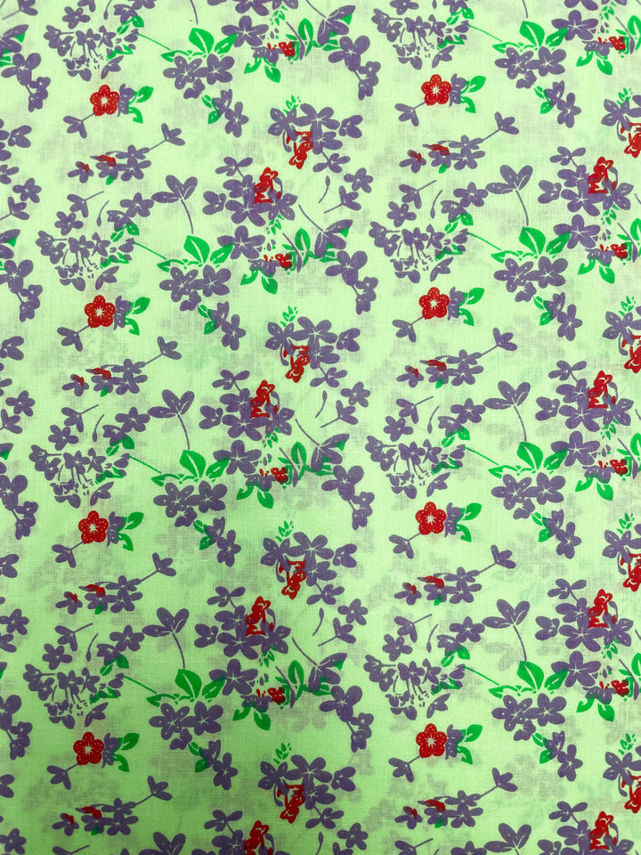 The Linen Voile - Jazzy by Super Cheap Fabrics is a 140cm wide fabric featuring a beautiful floral pattern with purple flowers and green leaves scattered across a light green background. Small red flowers are interspersed among the purple ones, creating a colorful and vibrant design that's perfect for home decor. This breathable fabric adds freshness to any space.