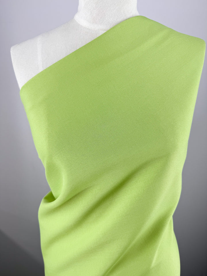 A mannequin displays Super Cheap Fabrics' Twill Suiting - Green Glow (150cm), a vibrant light green polyester with a diagonal weave, draped from chest to hips and wrapped diagonally across the chest and one shoulder.