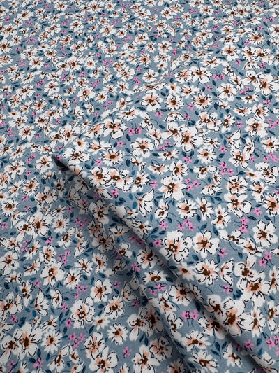 A close-up of Super Cheap Fabrics’ Cotton Sateen - Bluebell Meadows (150cm) shows neatly folded blue fabric with white, brown, and pink floral patterns—ideal for multi-use projects.
