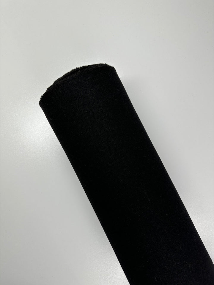 A roll of Super Cheap Fabrics' Wool Flannel in black, measuring 150cm wide, is positioned diagonally against a plain, light-gray background. The Italian wool appears smooth and tightly wrapped, with a slightly visible textured edge.