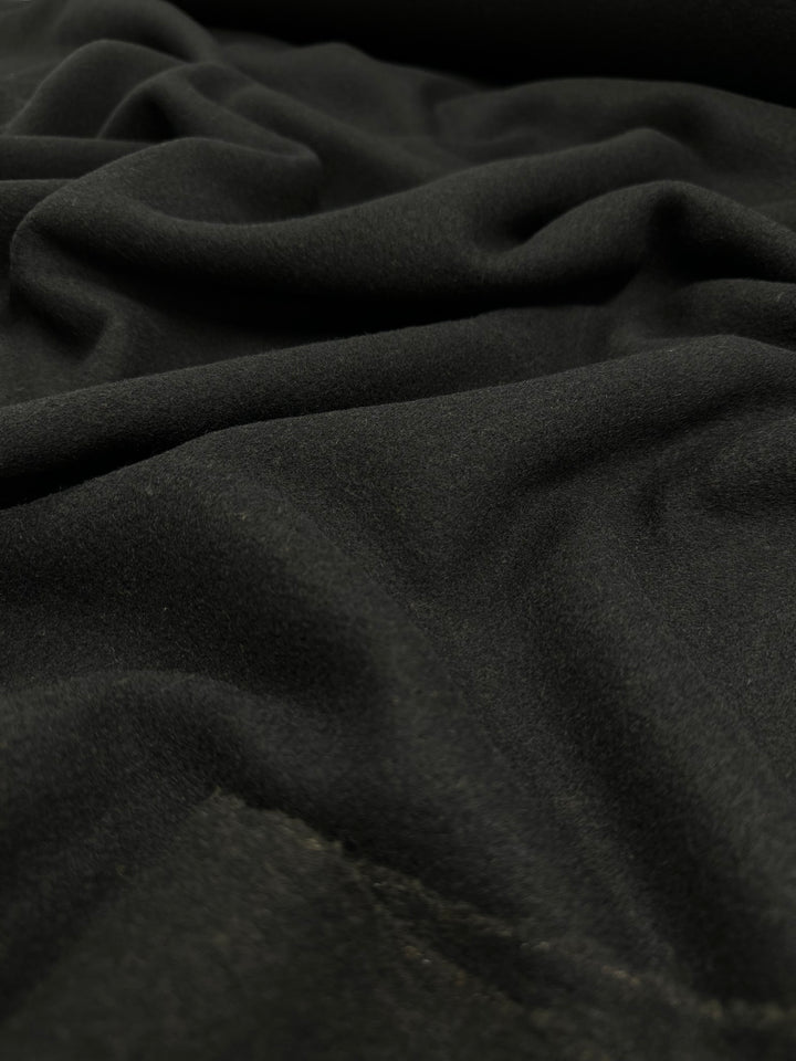 A close-up photograph of black Wool Flannel - Black - 150cm from Super Cheap Fabrics showcases its soft, velvety texture. The luxurious Italian wool is draped in gentle folds, creating smooth, flowing lines and subtle shadows across the surface. The material appears plush and opulent.