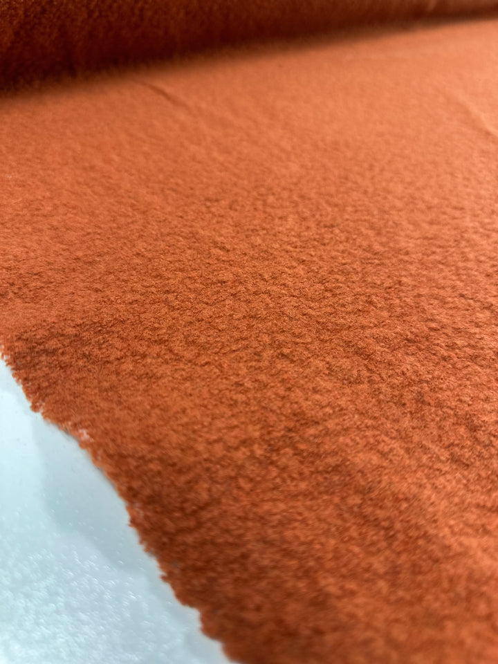 Close-up view of a roll of Super Cheap Fabrics' Wool Boucle in Burnt Orange - 135cm. The texture appears soft and slightly fuzzy, with the fabric unrolling over a smooth, light-colored surface. Resembling heavy-weight wool boucle fabric, the focus is on the details of the fabric’s surface.