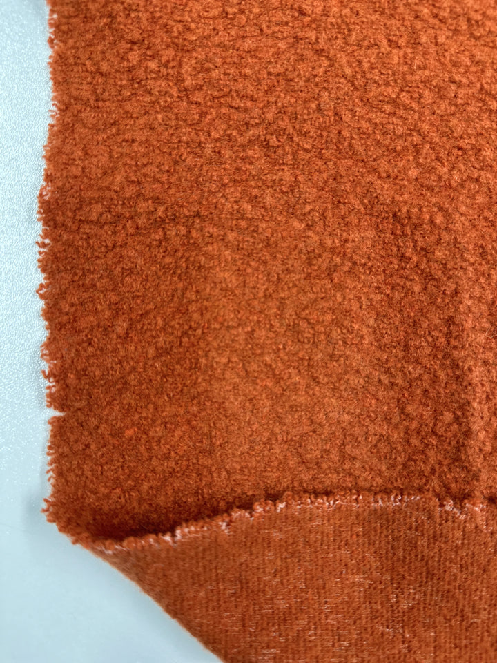 Close-up of a corner of Super Cheap Fabrics' Wool Boucle in Burnt Orange, measuring 135cm. The fabric features a heavy weight with a rough texture and a soft, fuzzy surface. The edges are slightly frayed, while the lower part is rolled up to reveal a smoother underside—perfect for crafting luxurious overcoats.