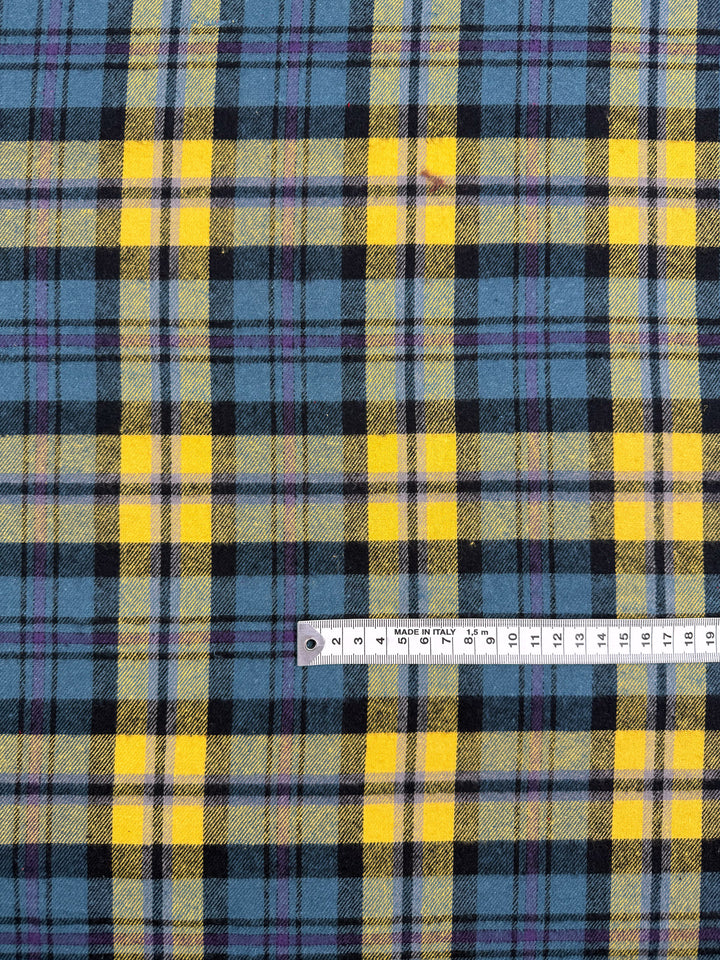 A close-up of Super Cheap Fabrics' Flannel Plaid - Highland Dusk (145cm), showing yellow, black, blue, and purple stripes with a metal centimeter ruler for scale; perfect medium weight fabric for shirts.