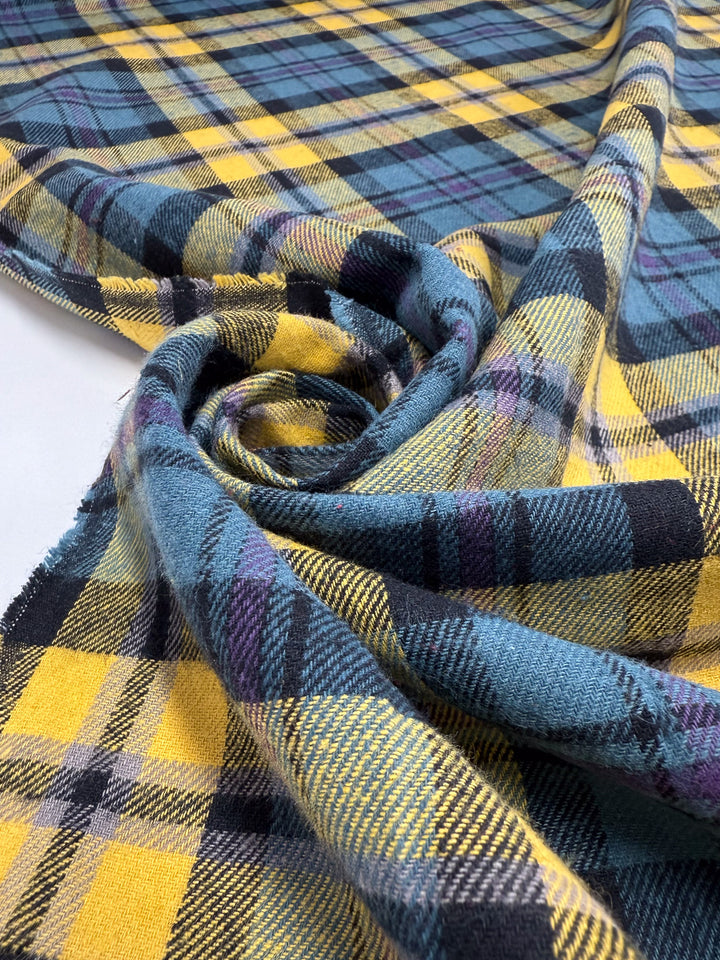 A close-up of Super Cheap Fabrics' Flannel Plaid - Highland Dusk (145cm), a medium weight cotton flannel in yellow, blue, black, and purple plaid. Soft-textured and loosely rolled on a white surface—ideal for shirts.
