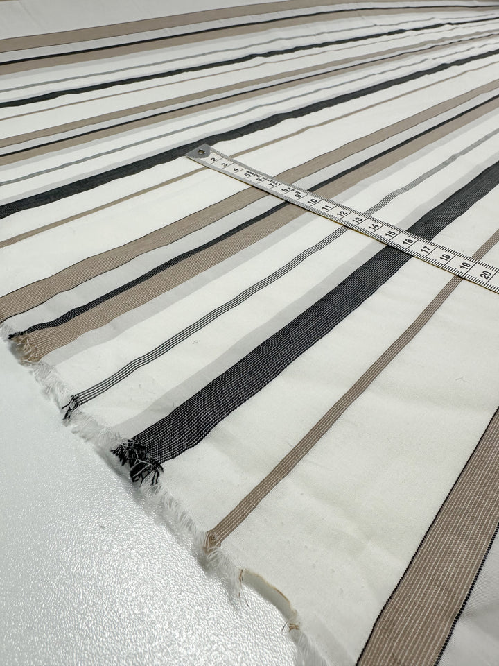 A close-up view of a piece of Super Cheap Fabrics' Cotton Voile - Creamy Nimbus - 155cm featuring horizontal stripes in black, brown, and white shades. A measuring tape is laid across the lightweight, breathable fabric, showing the scale. The edges are unfinished with small frays visible.