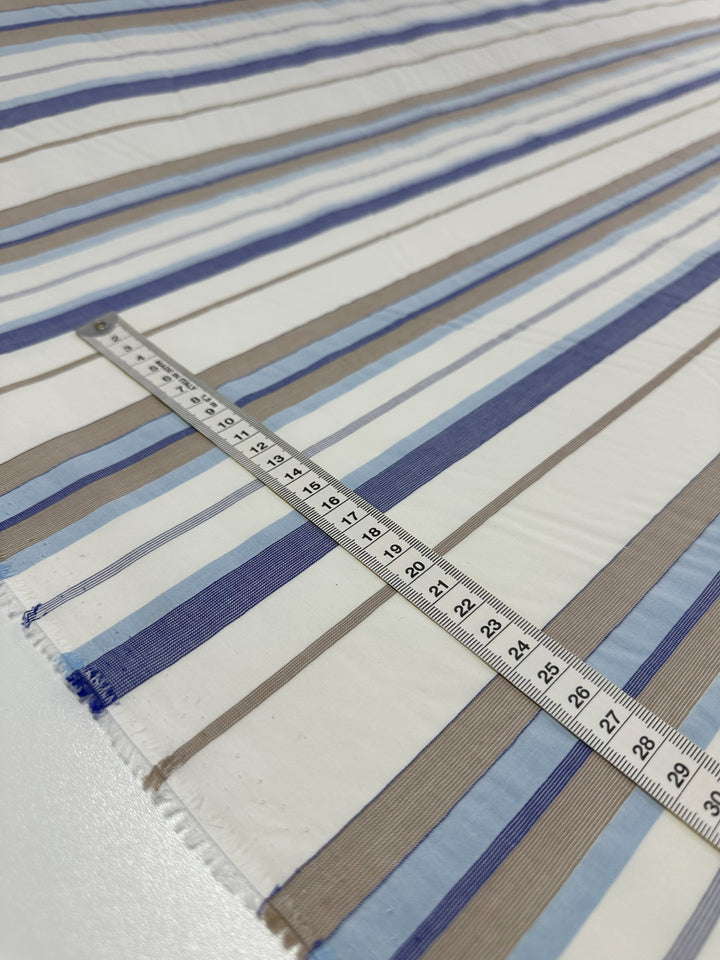 A piece of *Super Cheap Fabrics* Cotton Voile - Blue Breeze - 155cm, featuring alternating vertical stripes in shades of blue, brown, and white, lies flat on a surface. A measuring tape is placed horizontally across the fabric, displaying measurements in centimeters. Ideal for crafting light and airy garments.