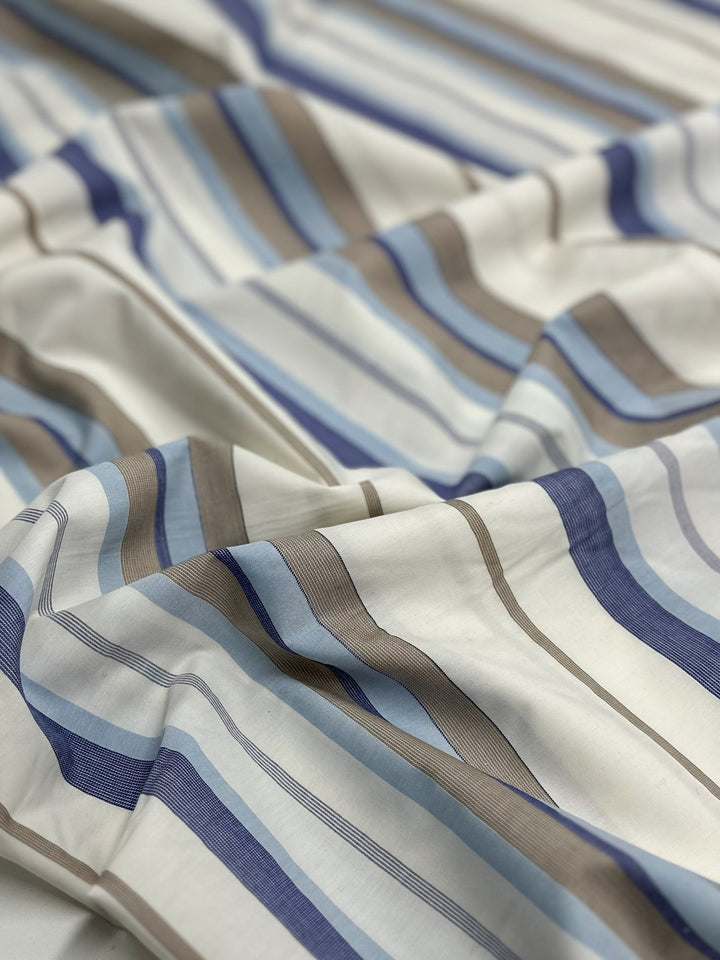 A close-up of Super Cheap Fabrics' Cotton Voile - Blue Breeze - 155cm showcases 100% cotton fabric with horizontal stripes in various shades of blue, brown, and white. The soft material appears slightly wrinkled, giving a casual and comfortable appearance. Ideal for light and airy garments, the stripes are evenly spaced and vary in thickness.