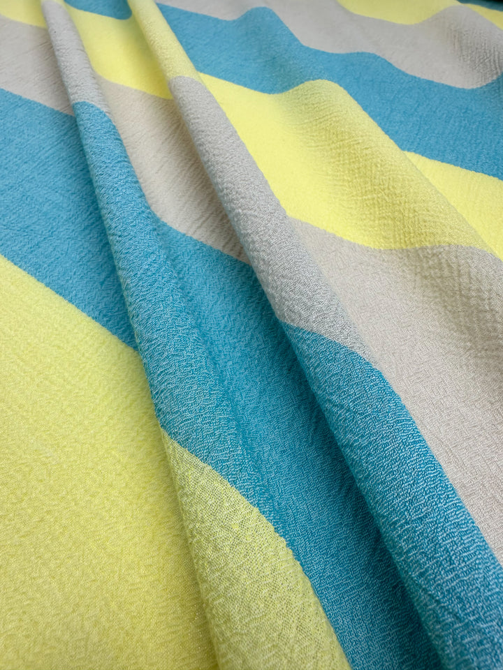 A close-up of Super Cheap Fabrics' Cotton Gauze - Beachy - 130cm reveals lightweight cotton gauze with wide, horizontal stripes in bright yellow, beige, and turquoise. The fabric's slightly grainy texture makes it perfect for home decor or children's clothing due to its soft and lightweight nature with gentle folds.