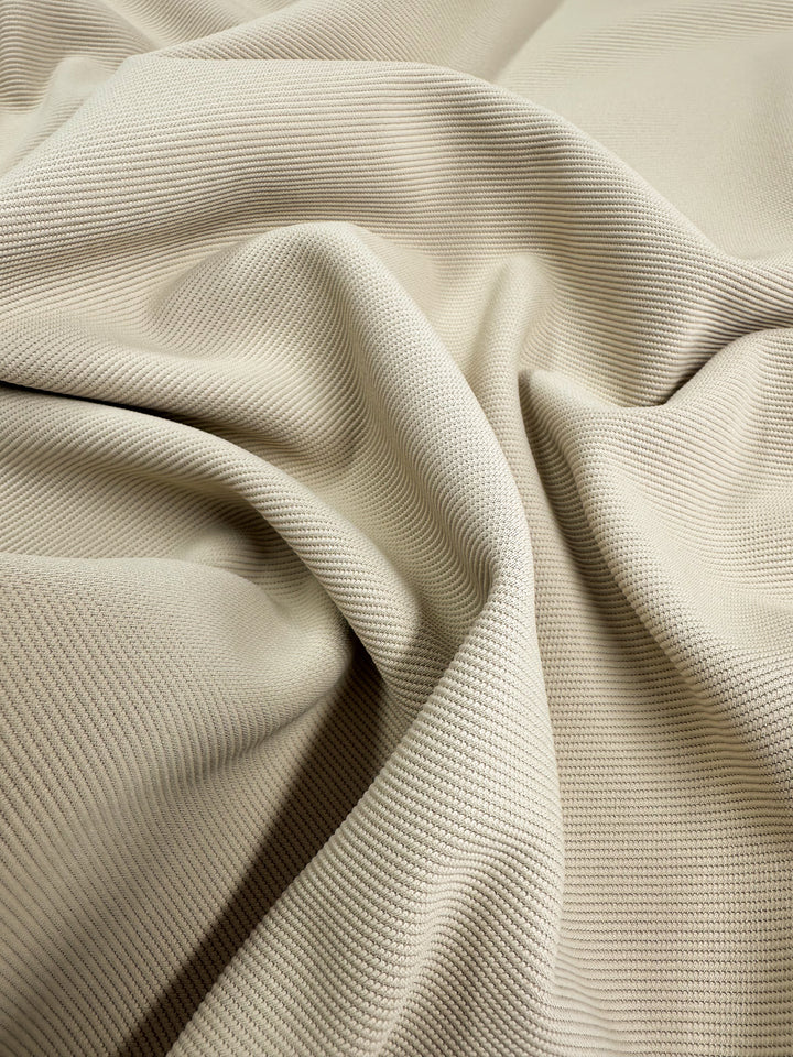 A close-up image of Super Cheap Fabrics' Stretch Twill in Putty, available in a 135cm width. The beige material features a textured knit fabric with a wavy, folded surface and a subtle striped pattern that adds depth and visual interest, making it soft to the touch.