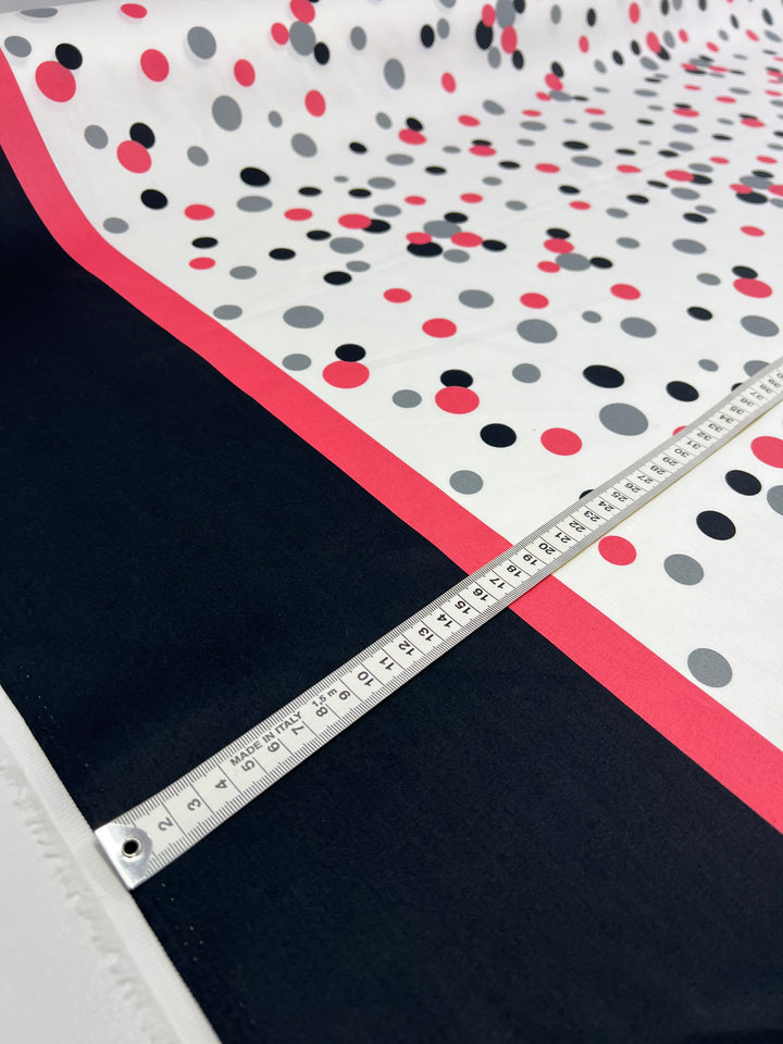 A piece of Cotton Sateen - Bordered Bubbles - 145cm from Super Cheap Fabrics, featuring a 100% cotton fabric with black, red, and gray polka dots and a black and red border, is displayed. A measuring tape laid across this lightweight material shows measurements in centimeters, indicating the fabric's size—ideal for household décor items.