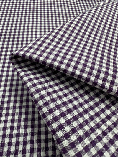 A close-up image of Cotton Gingham - Mini Purple - 140cm by Super Cheap Fabrics, made from 100% cotton. The fabric shows a repetitive checkered pattern with small, evenly spaced squares. A portion of the lightweight fabric is folded, highlighting its texture and flexibility.