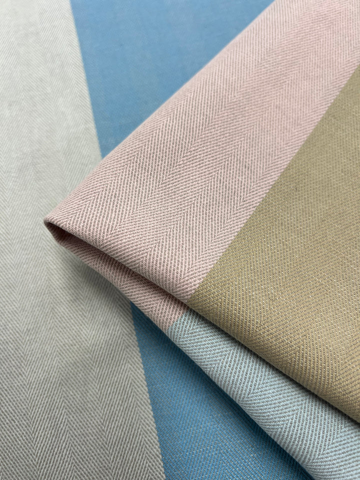 Close-up of folded fabric with a herringbone pattern from the Designer Cotton - Blipe - 155cm by Super Cheap Fabrics. The fabric features wide vertical stripes in pastel colors, including light pink, beige, blue, and grey, crafted using sustainable deadstock materials for eco-friendly fashion.