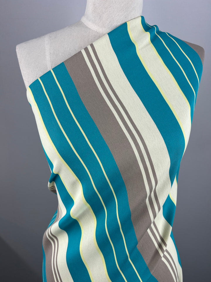 A mannequin is draped with a fabric from our Designer Cotton - St.Tropez - 157cm by Super Cheap Fabrics, featuring a bold, striped pattern in shades of teal, gray, white, and light yellow. Crafted from sustainable deadstock materials, the fabric is wrapped diagonally across the torso for a visually striking and eco-friendly look.