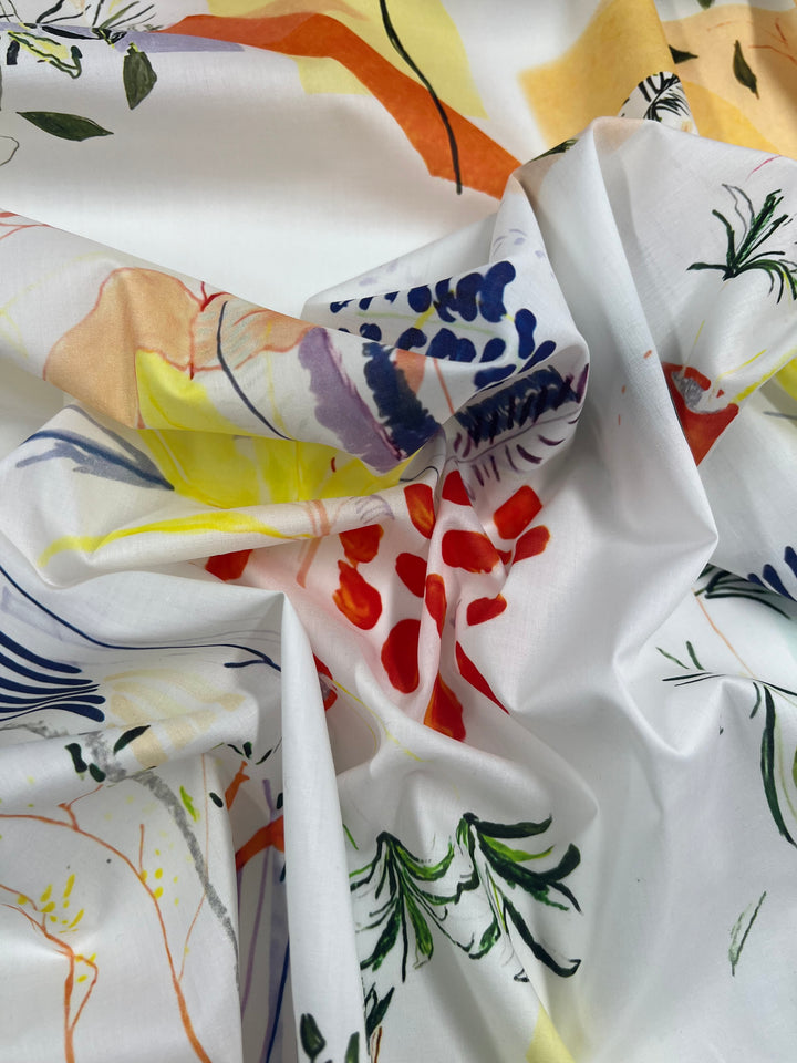 A piece of crumpled Designer Cotton - Holiday - 136cm fabric from Super Cheap Fabrics with a colorful floral and abstract design. The patterns feature splashes of red, yellow, and purple, along with green leaves against a white background. This lightweight fabric is perfect for multi-use applications.